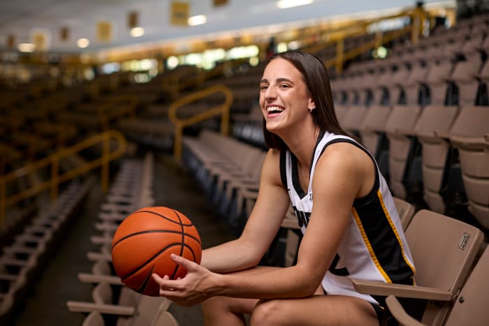 Caitlin Clark smiles and holds a basketball while posing for a portrait in the stands.